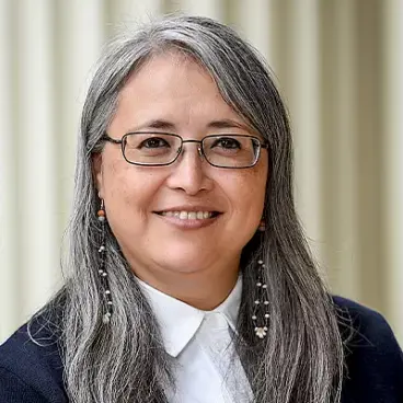 A person with long gray hair, glasses, and dangling earrings smiles while wearing a white collared shirt and dark jacket. The background features vertical light-colored lines.