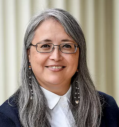 A person with long gray hair, glasses, and dangling earrings smiles while wearing a white collared shirt and dark jacket. The background features vertical light-colored lines.