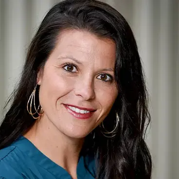 A woman with long dark hair and hoop earrings smiles at the camera. She is wearing a teal top, and the background features light vertical drapes.