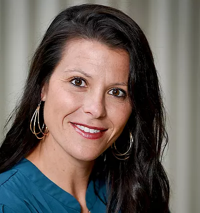 A woman with long dark hair and hoop earrings smiles at the camera. She is wearing a teal top, and the background features light vertical drapes.