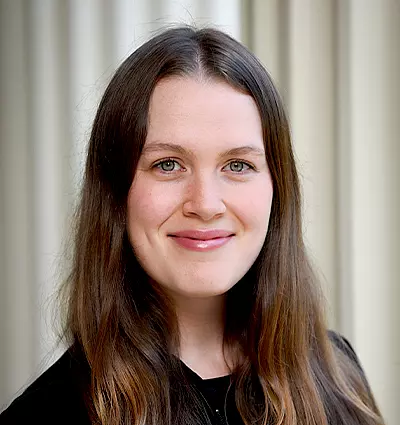 Portrait of a smiling person with long brown hair, wearing a black top. They are standing in front of a light-colored, softly blurred background.