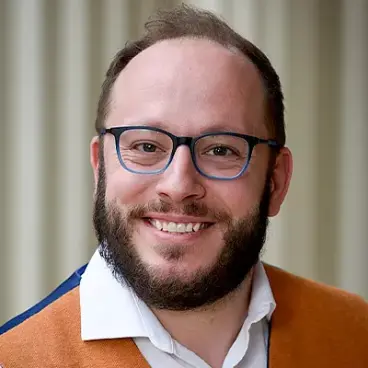 Smiling man with glasses and a beard, wearing a white shirt and an orange vest, standing in front of a blurred background.