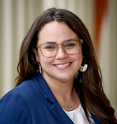 A person with long brown hair and glasses is smiling at the camera. They are wearing a blue blazer, a light-colored blouse, and drop earrings. The background is softly blurred, suggesting an outdoor setting.