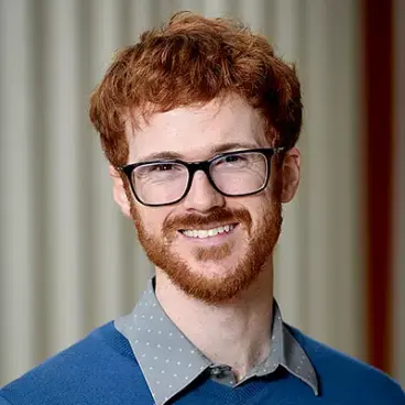 A person with red hair and a beard, wearing glasses and a blue sweater over a gray shirt, smiles at the camera. The background is softly blurred with vertical stripes.