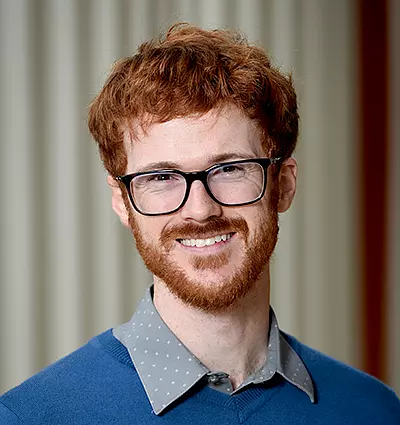 A person with red hair and a beard, wearing glasses and a blue sweater over a gray shirt, smiles at the camera. The background is softly blurred with vertical stripes.