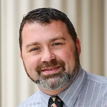 A person with short dark hair and a beard is smiling. They are wearing a striped shirt and a tie with a pattern. The background features blurred vertical lines.