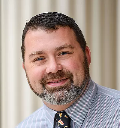 A person with short dark hair and a beard is smiling. They are wearing a striped shirt and a tie with a pattern. The background features blurred vertical lines.