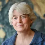 A person with short gray hair is smiling softly, wearing a textured blue jacket. The background is an abstract, blurred pattern with warm tones.