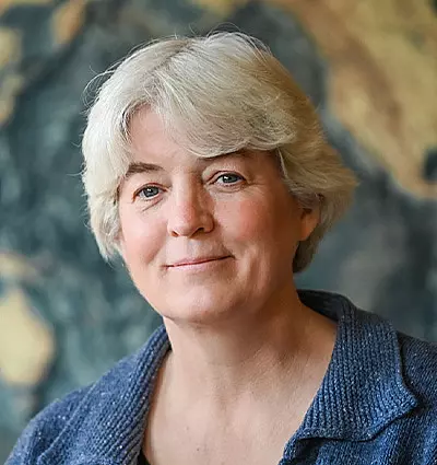 A person with short gray hair is smiling softly, wearing a textured blue jacket. The background is an abstract, blurred pattern with warm tones.