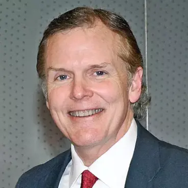 A middle-aged man in a suit and red tie smiles at the camera. The background is a gray, perforated wall.