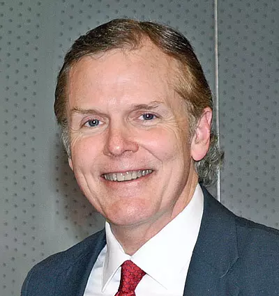 A middle-aged man in a suit and red tie smiles at the camera. The background is a gray, perforated wall.