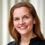 A woman with long brown hair is smiling at the camera. She is wearing a black top and pink flower-shaped earrings. The background is softly blurred, with vertical stripes.
