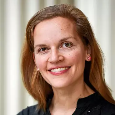 A woman with long brown hair is smiling at the camera. She is wearing a black top and pink flower-shaped earrings. The background is softly blurred, with vertical stripes.