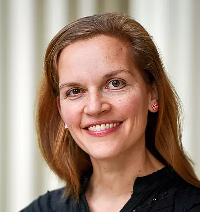 A woman with long brown hair is smiling at the camera. She is wearing a black top and pink flower-shaped earrings. The background is softly blurred, with vertical stripes.
