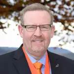 A man wearing glasses, a black suit, and an orange tie is smiling. He has short hair and a trimmed beard. The background features blurred autumn leaves and a clear sky.