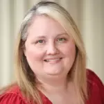 A person with shoulder-length blonde hair smiles at the camera. They are wearing a red top. The background is softly blurred.