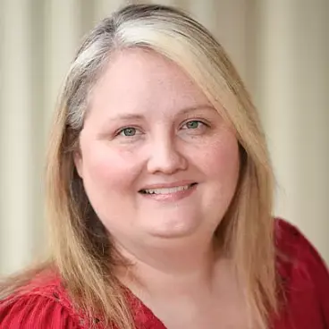A person with shoulder-length blonde hair smiles at the camera. They are wearing a red top. The background is softly blurred.