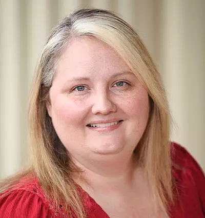 A person with shoulder-length blonde hair smiles at the camera. They are wearing a red top. The background is softly blurred.