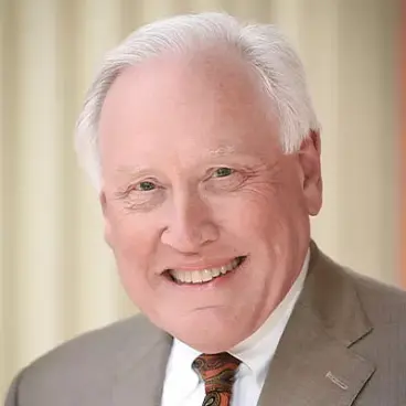 A smiling older man with white hair is wearing a brown suit, white shirt, and a patterned tie. The background is softly blurred with vertical lines.