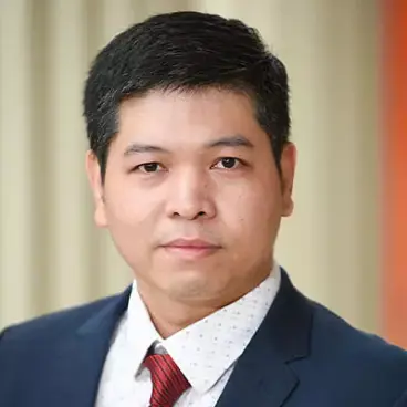 A person with short dark hair is wearing a dark suit, white shirt, and red tie. They have a neutral expression and are set against a blurred background.