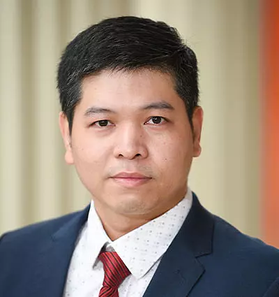 A person with short dark hair is wearing a dark suit, white shirt, and red tie. They have a neutral expression and are set against a blurred background.