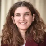 Woman with long, wavy brown hair, smiling. She's wearing a burgundy jacket and a light-colored top. The background is softly blurred with neutral tones.