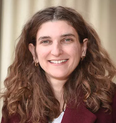 Woman with long, wavy brown hair, smiling. She's wearing a burgundy jacket and a light-colored top. The background is softly blurred with neutral tones.
