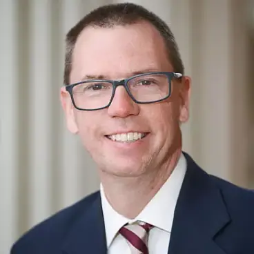 A smiling man with short hair, wearing glasses and a formal suit with a striped tie. The background is softly blurred, emphasizing his professional appearance.