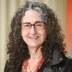 A person with curly gray hair and glasses smiles at the camera. They are wearing a black jacket over a coral shirt with a floral pattern. The background is softly blurred.