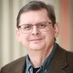 A middle-aged man with glasses smiles slightly. He is wearing a brown herringbone jacket over a gray shirt. The background is softly blurred with beige and orange tones.