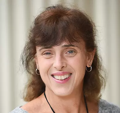 A person with brown hair smiles, wearing hoop earrings and a gray top. The background is softly blurred and neutral in color.