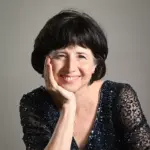 Woman with short dark hair and a warm smile, wearing a sparkly dark blouse. She rests her head on her hand, set against a plain background.