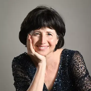 Woman with short dark hair and a warm smile, wearing a sparkly dark blouse. She rests her head on her hand, set against a plain background.