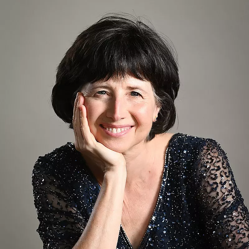 Woman with short dark hair and a warm smile, wearing a sparkly dark blouse. She rests her head on her hand, set against a plain background.