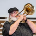 A bearded man wearing glasses and a black cap plays a brass trombone. He is dressed in a dark shirt and stands in front of a blurred background, concentrating on his performance.