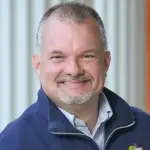 A man with short hair and a beard smiles at the camera. He is wearing a navy blue jacket over a light-colored shirt. The background features vertical white and orange stripes.
