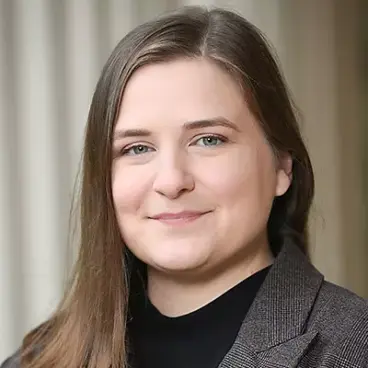 A person with long brown hair is smiling softly, wearing a dark top and a gray blazer. The background is softly blurred with vertical lines.