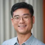 A person with short black hair, wearing round glasses and a light gray collared shirt, smiles warmly in front of a blurred, striped background.