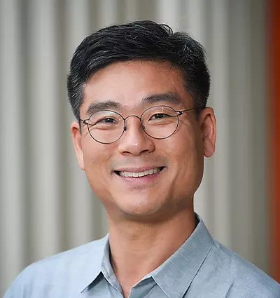 A person with short black hair, wearing round glasses and a light gray collared shirt, smiles warmly in front of a blurred, striped background.