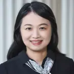 A smiling woman with long dark hair wearing a black top and a patterned scarf, standing against a light-colored background featuring vertical lines.
