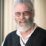 A smiling man with white hair and a beard wears round glasses, a dark vest, and a white shirt. The background is softly blurred with vertical lines.