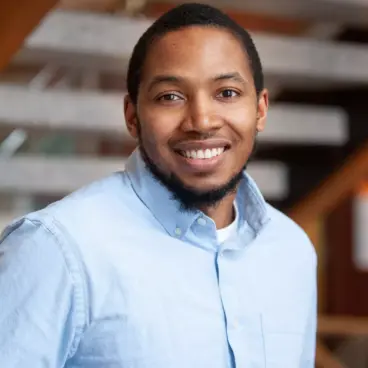 A smiling person wearing a light blue button-up shirt stands indoors against a blurred background of wooden beams and metal railings.