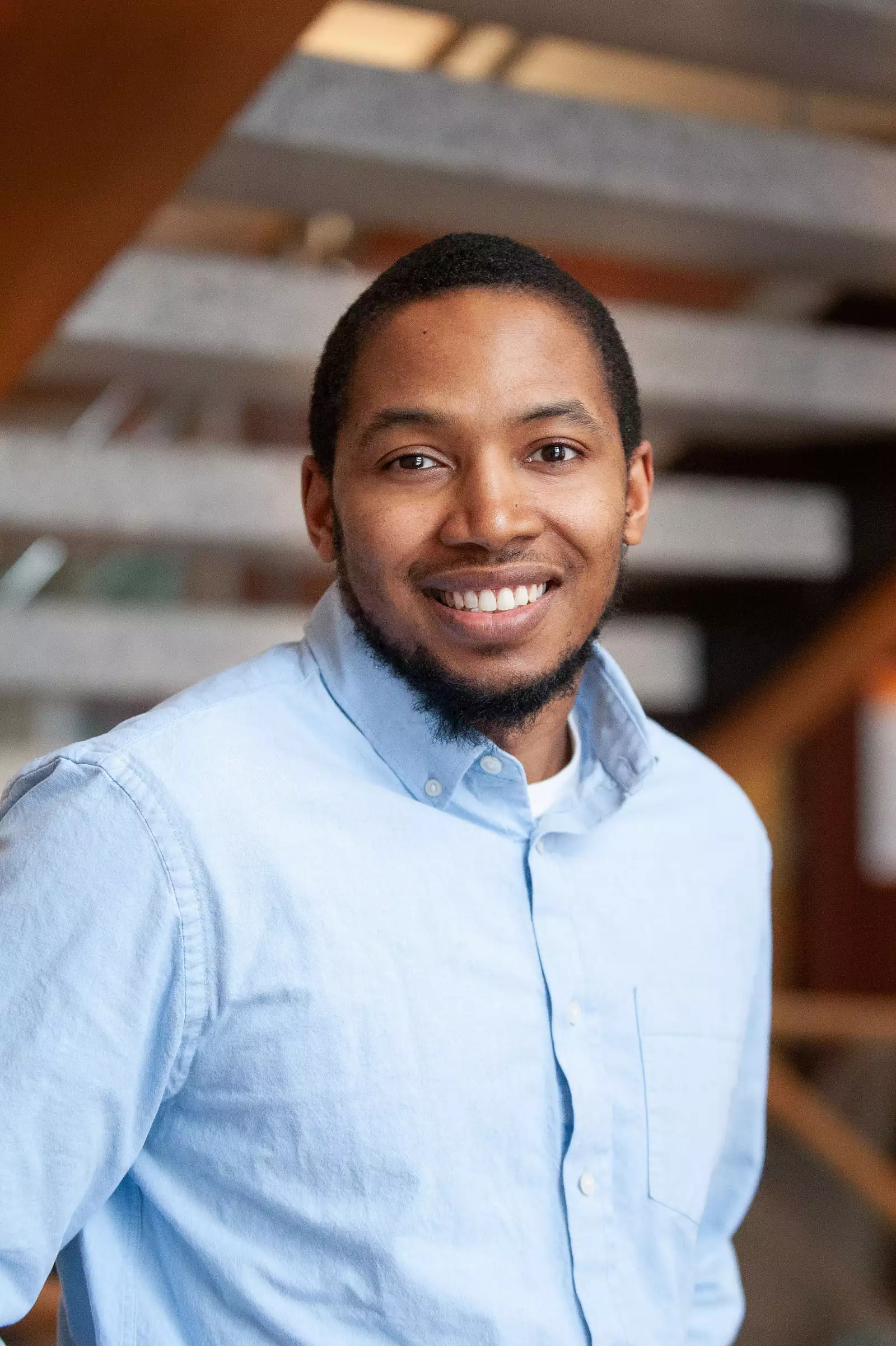 A smiling person wearing a light blue button-up shirt stands indoors against a blurred background of wooden beams and metal railings.