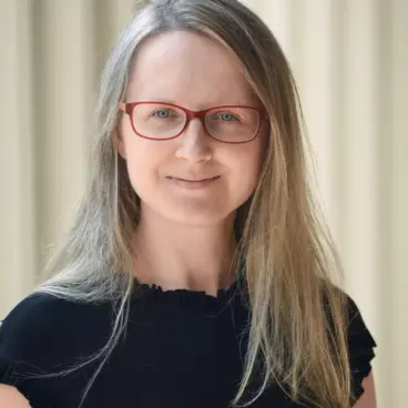 A person with long blonde hair and red glasses is smiling softly while wearing a black top. The background is blurred vertical beige stripes.