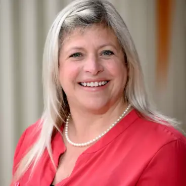 Smiling woman with long, light-colored hair wearing a red blouse and pearl necklace, standing in front of a light-colored curtain.