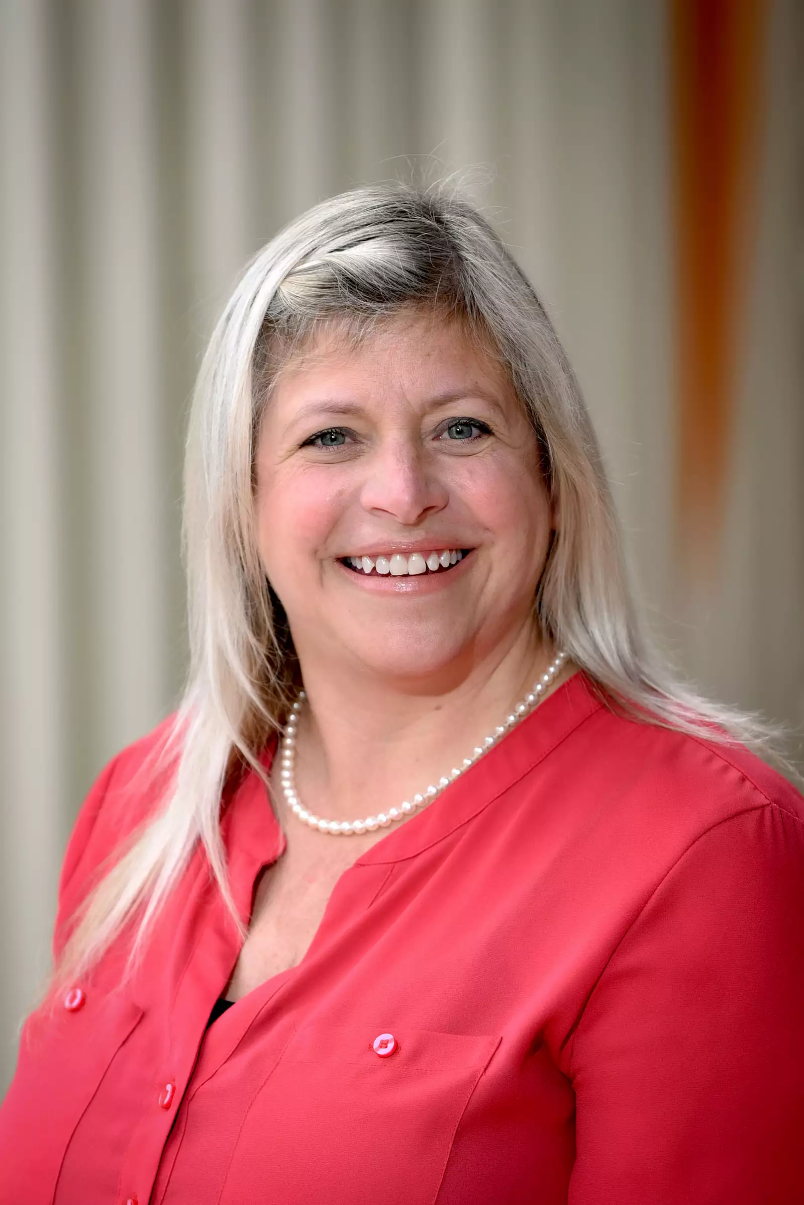 Smiling woman with long, light-colored hair wearing a red blouse and pearl necklace, standing in front of a light-colored curtain.