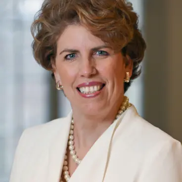 A woman with short, wavy hair smiles warmly. She wears a white blazer, pearl earrings, and a matching pearl necklace. The background is softly blurred, suggesting an indoor setting.