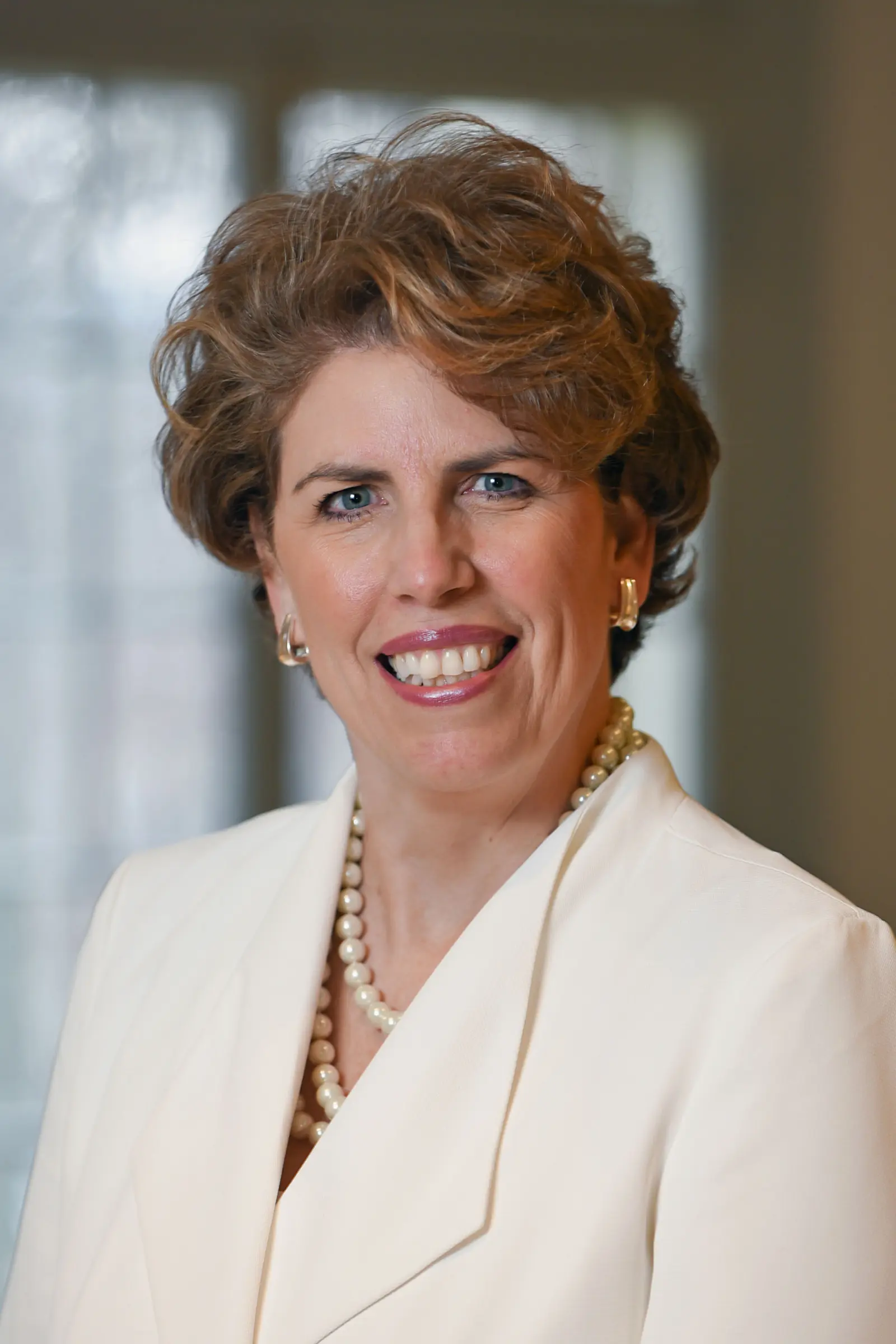 A woman with short, wavy hair smiles warmly. She wears a white blazer, pearl earrings, and a matching pearl necklace. The background is softly blurred, suggesting an indoor setting.