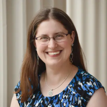 A woman with long brown hair and glasses smiles warmly. She is wearing a blue and black patterned top and a necklace with a circular pendant. The backdrop features neutral-colored vertical panels.