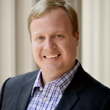 A person with short blond hair is smiling, wearing a dark jacket over a plaid shirt. The background is softly blurred, featuring vertical lines.